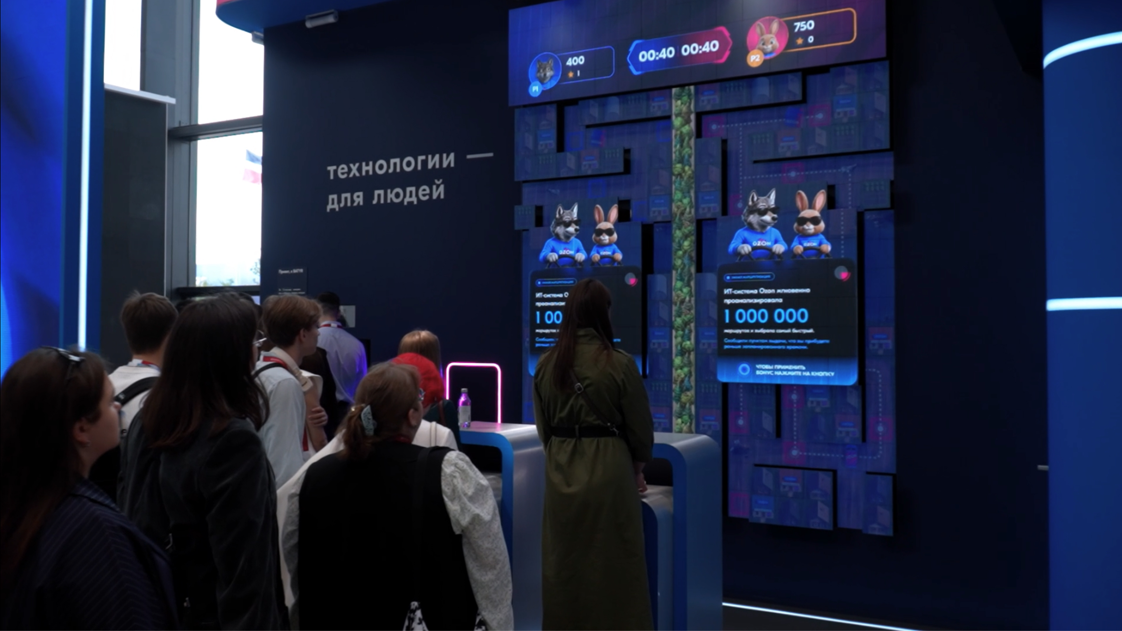 Стенд Ozon: гости у стоек с джойстиками, на кинетическом экране — уровень с деревнями и плашка «Умная маршрутизация»