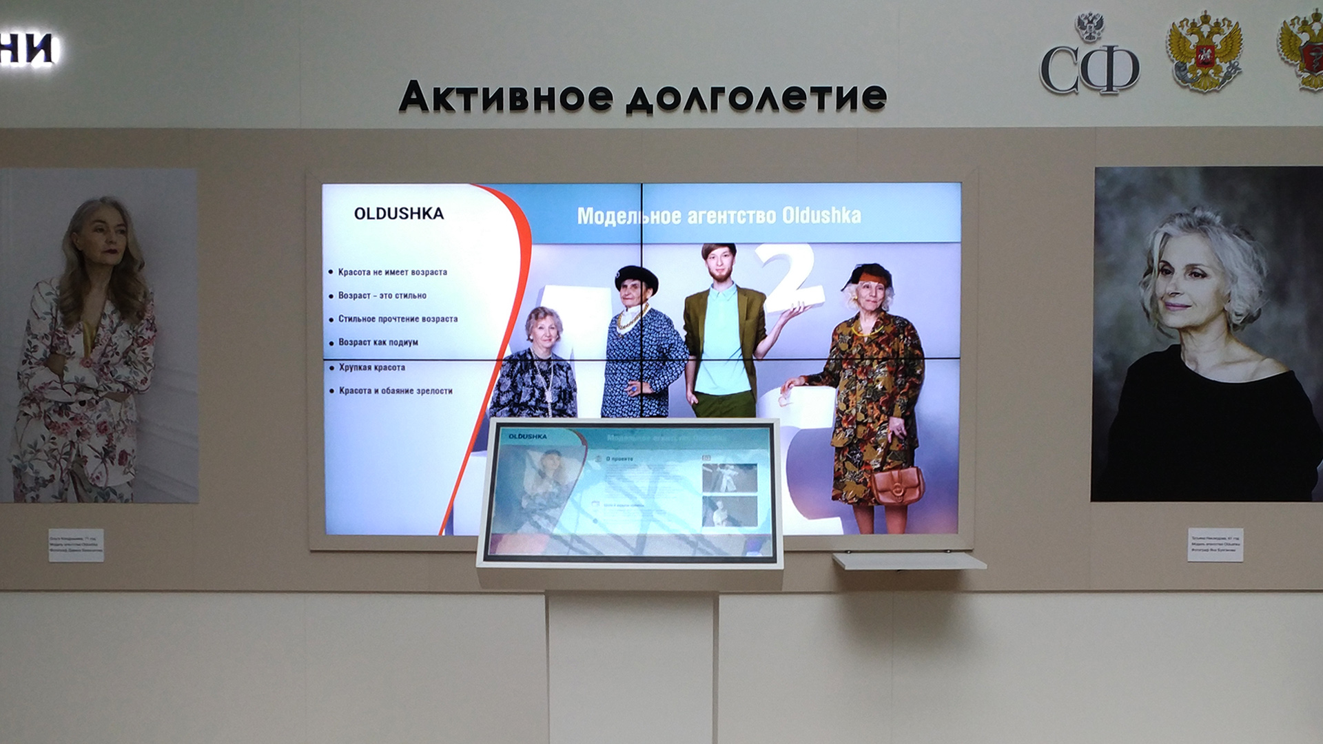 Стена с надписью «Активное долголетие»: видеостена с презентацией модельного агентства Oldushka и сенсорный экран под ней, по бокам — портреты