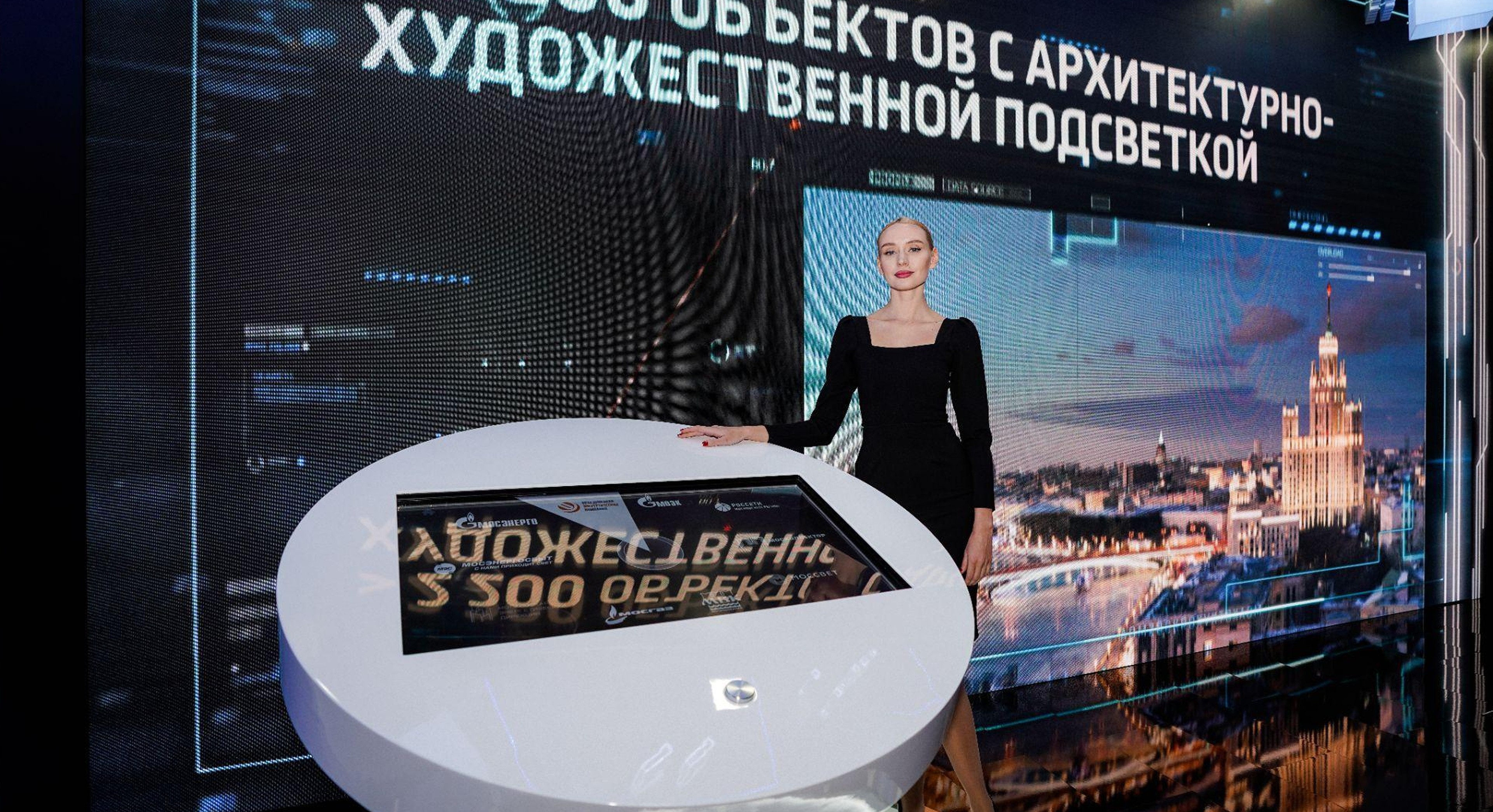 Большой экран с роликом об объектах с архитектурно-художественной подсветкой и пульт — круглый белый стол со встроенным экраном выбора компаний и металлической ручкой