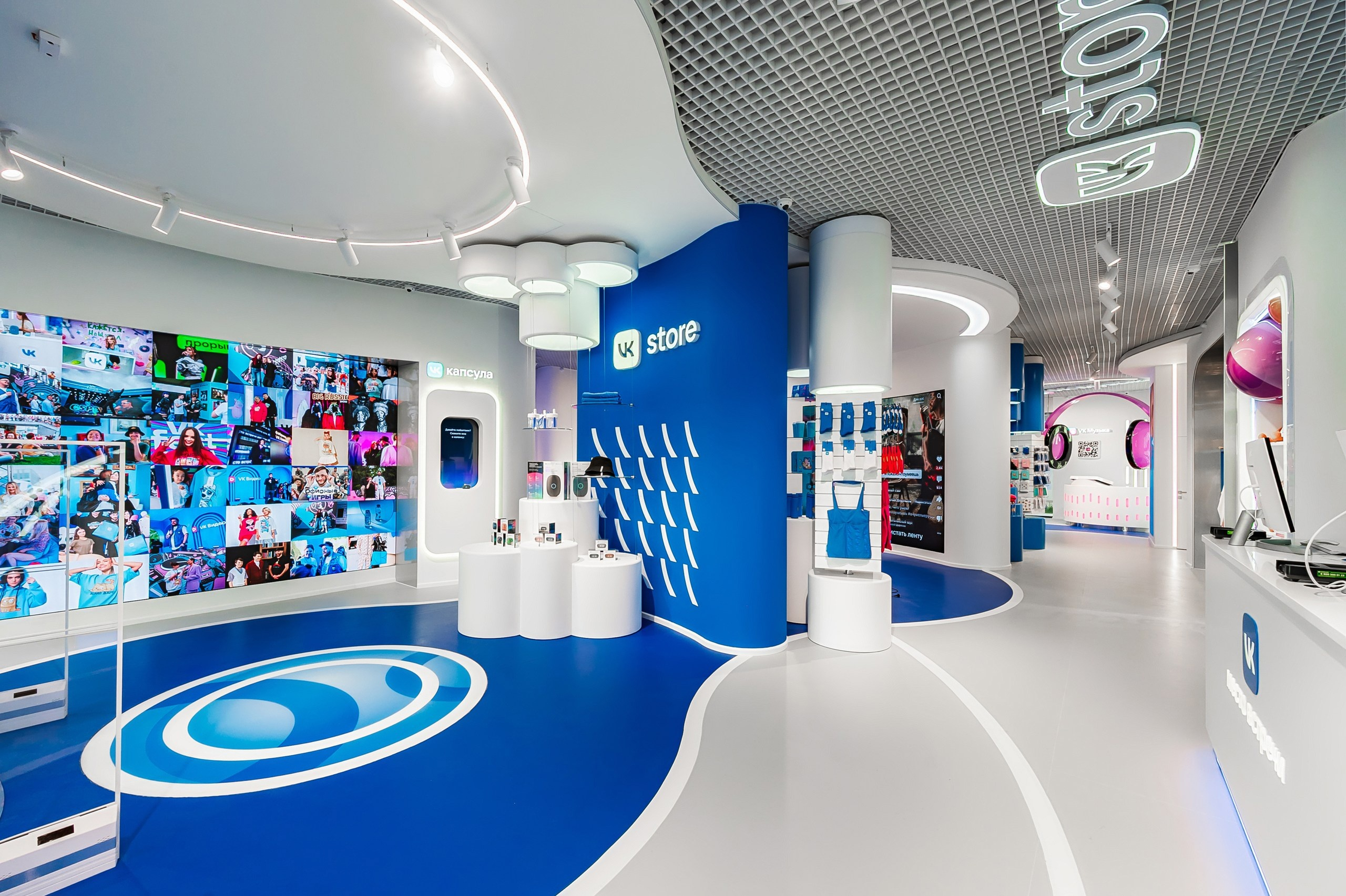 The sales floor: a blue floor with white paths, a video wall with a photo mosaic and a “VK Capsule” stand on the left, a blue wall with the words “VK store” in the centre, and pink headphones above the VK Music console at the back of the hall