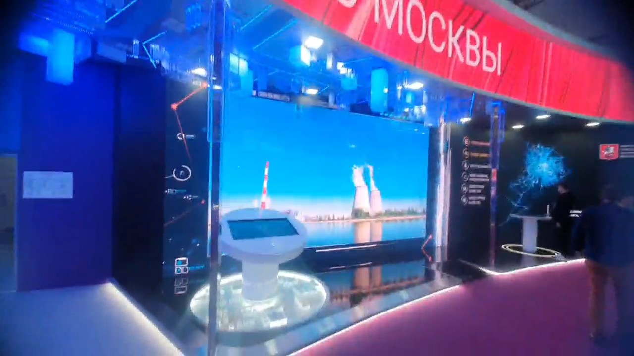 Стенд со стороны прохода: над входом красный светодиодный фриз со словом «Москвы», в центре большой экран с градирнями электростанции и круглый белый пульт перед ним, справа — тёмный экран с картой Москвы и круглым столом