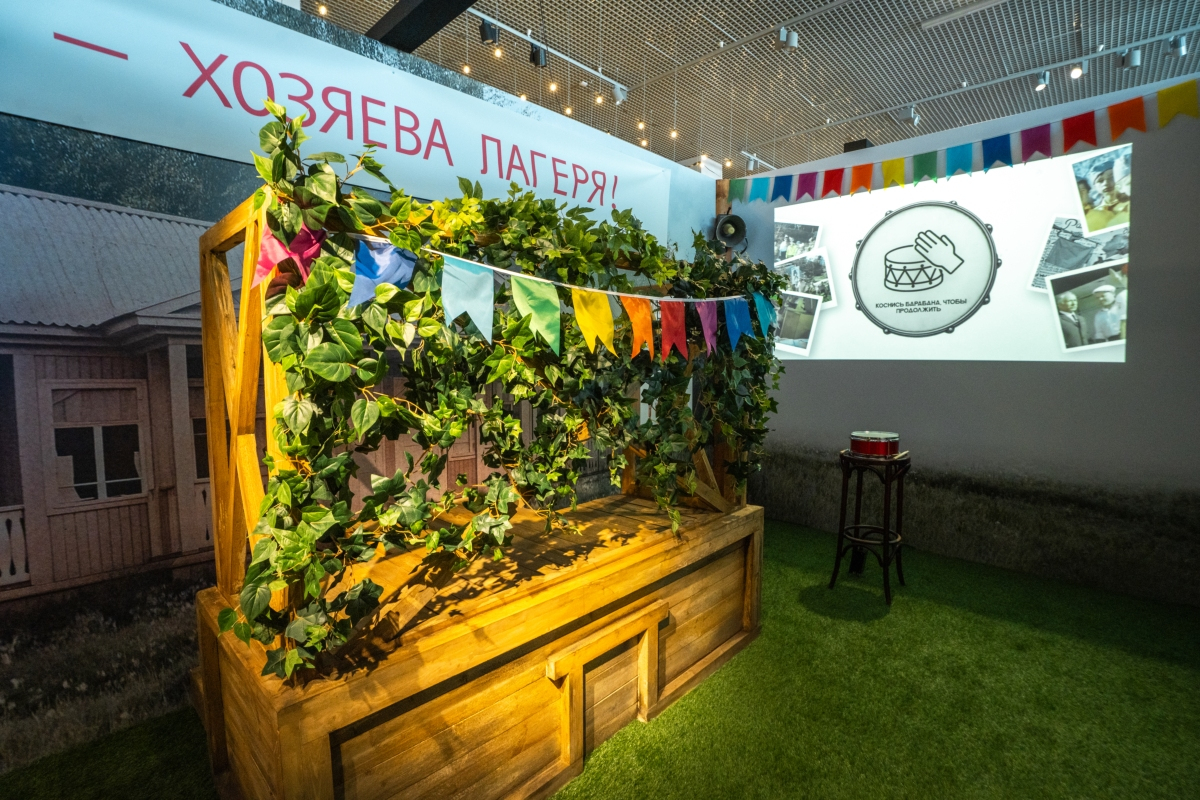 Сцена «Хозяева лагеря!», справа проекция с заставкой «Коснись барабана, чтобы продолжить», над ней громкоговоритель, барабан на табурете