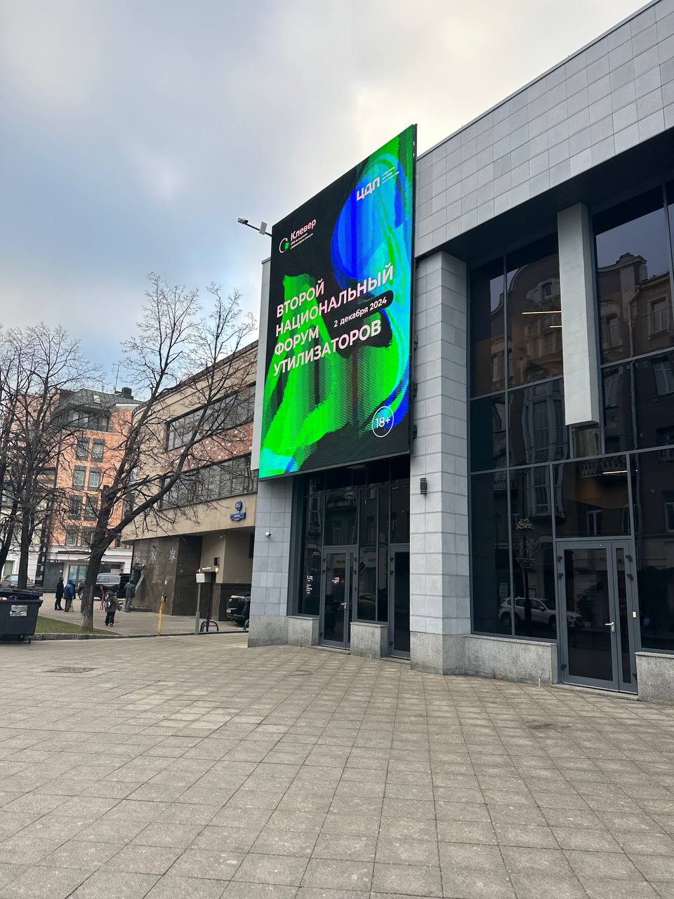 Corner of the DBS building from the street: a vertical media facade with the forum intro above the glass entrance, with bare trees and neighbouring buildings nearby