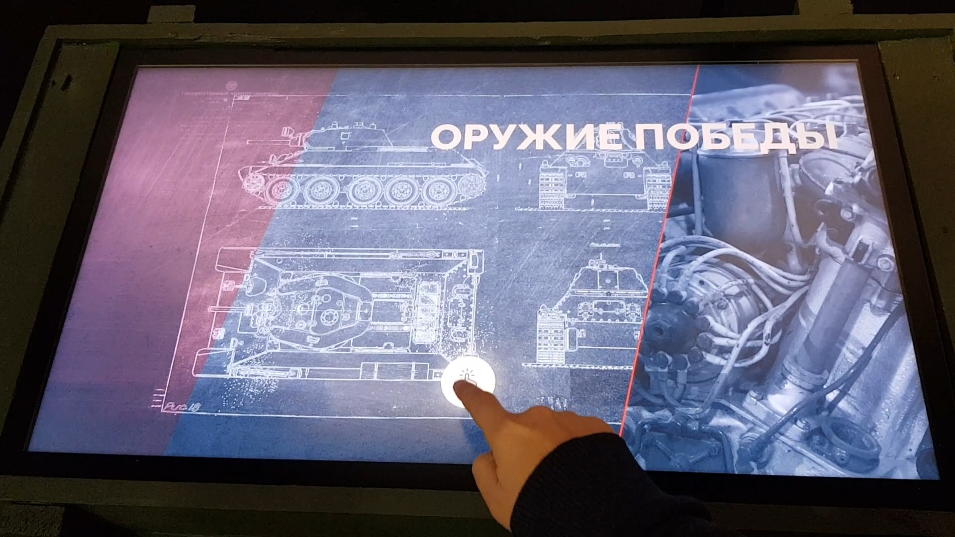 Экран в крышке зелёного оружейного ящика: чертежи танка и надпись «Оружие Победы», палец посетителя на кнопке запуска