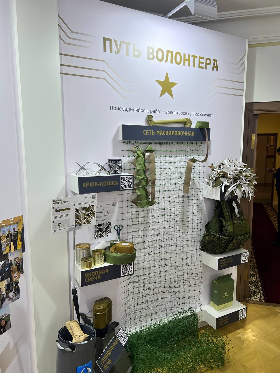 “A volunteer’s path” stand: the call “Join the volunteers’ work right now!”, a camouflage net, a grappling hook, trench candles, a chest rig and other objects on shelves with labels and QR codes