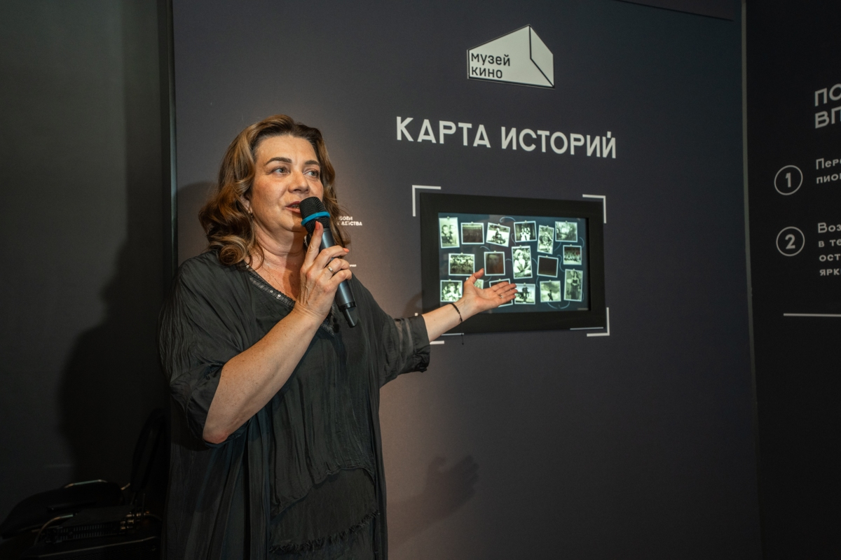 Выступающая с микрофоном на открытии выставки показывает на экран «Карты историй» с фотографиями