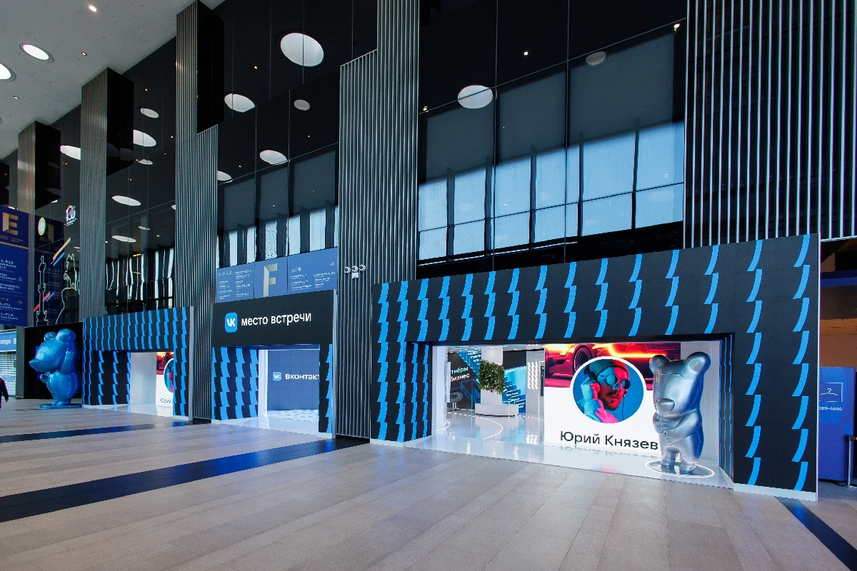 The long facade of the VK stand along the Expoforum hall: dark walls with a blue pattern, the “VK Meeting Point” sign, figures of a blue and a silver bear at the entrances, and a glowing screen with a portrait in the opening