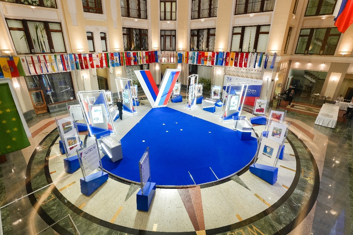 A view from above of the exhibition in the CEC atrium: a round blue podium with the “tick” in the colours of the tricolour in the centre, and around it illuminated frames with vertical touch screens, sloping touch tables, display cases and poster stands; flags of the regions above the hall