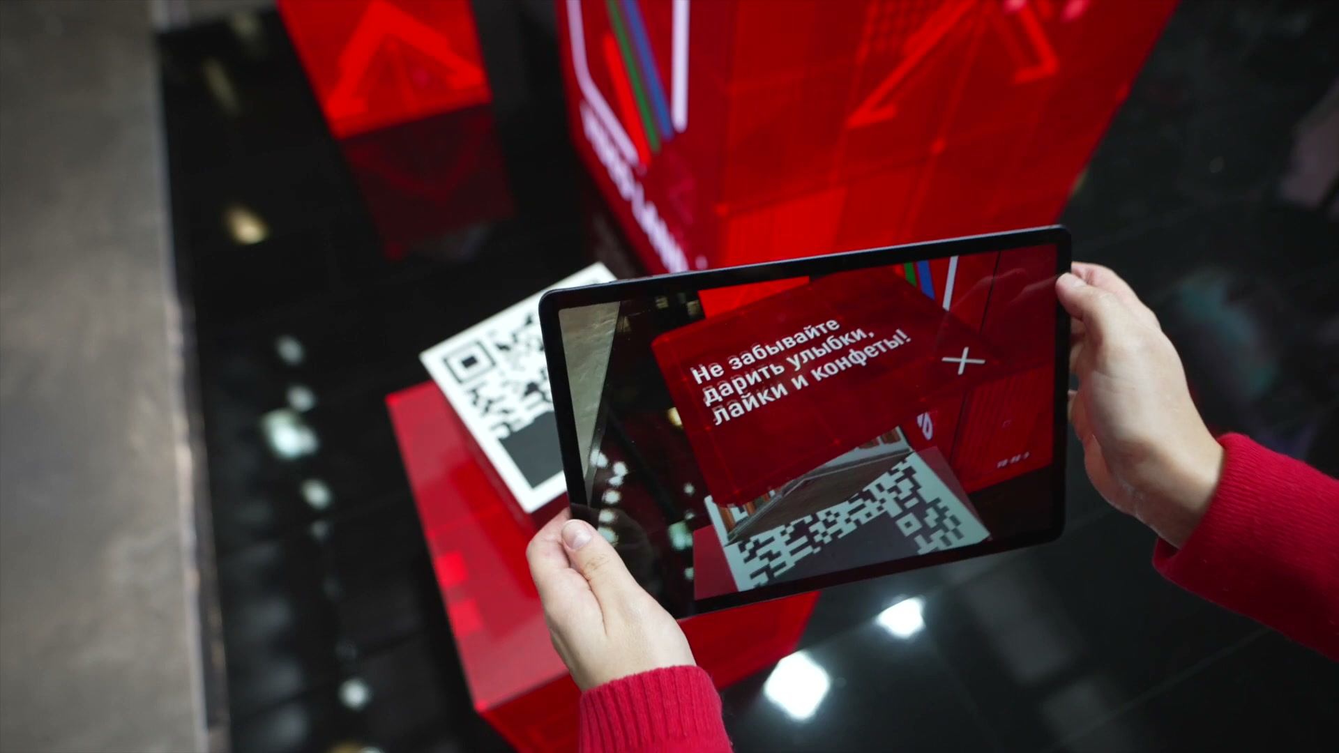 Руки держат планшет над красным кубом с QR-меткой, на экране — предсказание «Не забывайте дарить улыбки, лайки и конфеты!»