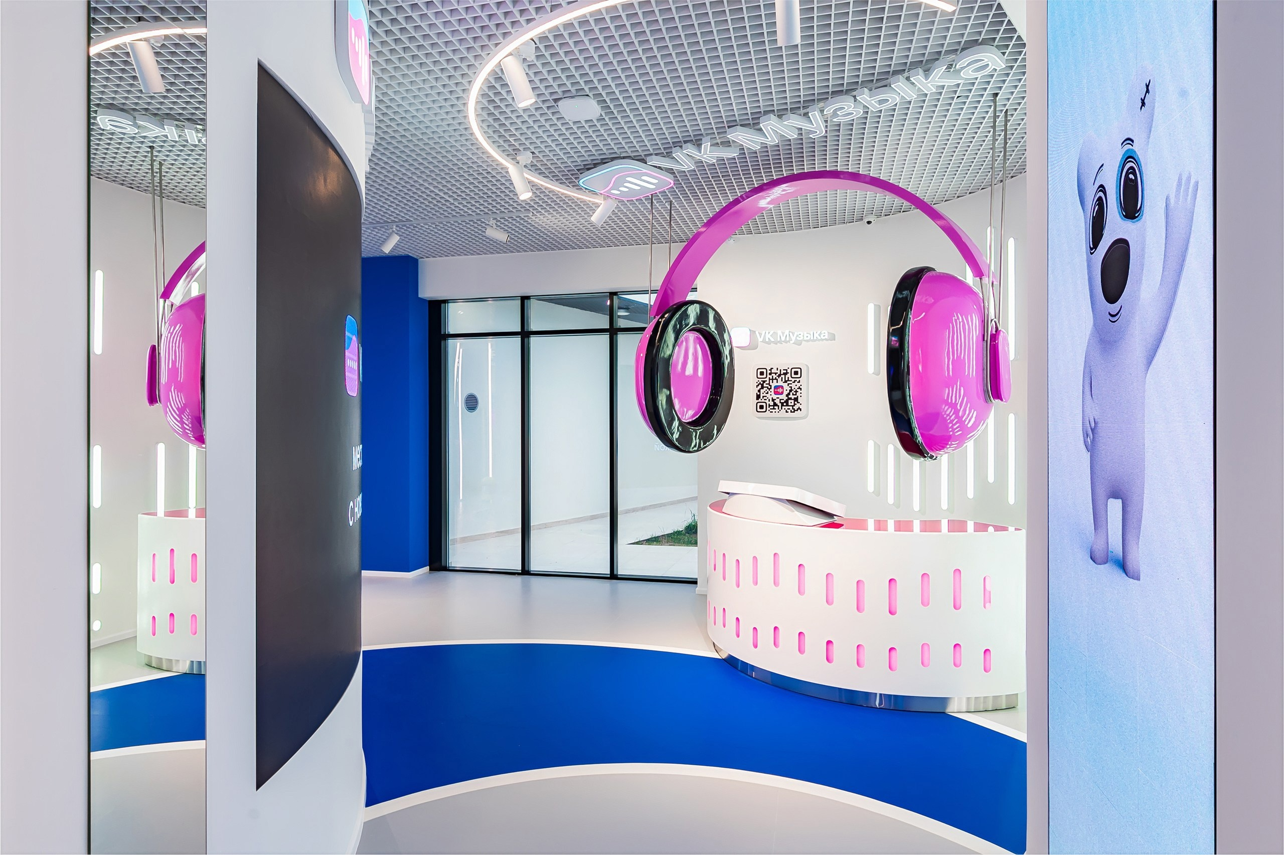 The VK Music zone: a white console table with giant pink headphones suspended above it, the words “VK Music” and a QR code on the wall, and a VK sticker character on the screen on the right