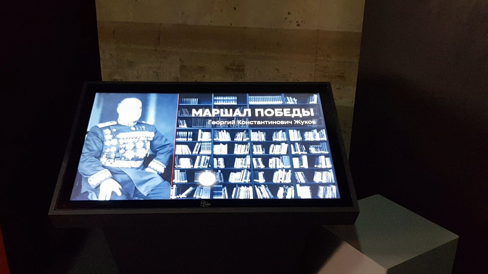 Наклонный сенсорный экран на подставке в затемнённом зале; на экране Жуков в парадном мундире и надпись «Маршал Победы»
