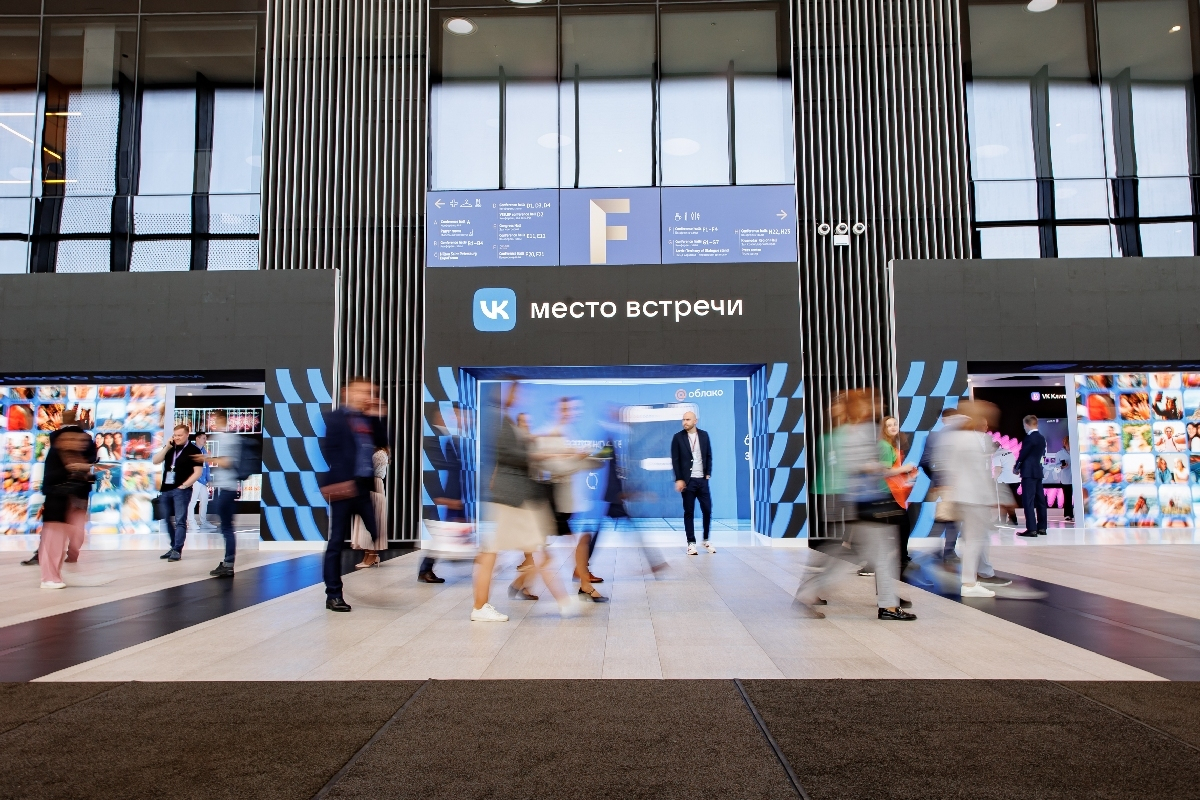 The entrance to the stand under the “VK Meeting Point” sign; forum guests walk past in motion, with walls of screens showing photos on either side of the entrance