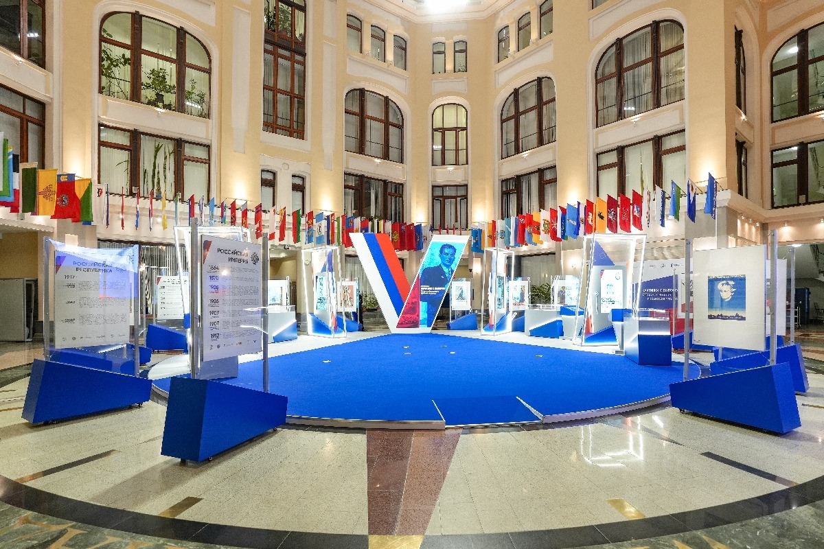 The exhibition in the CEC atrium at eye level: stands with a chronology of the eras in front, the “tick” with a portrait on its screen behind the blue podium, illuminated frames with vertical touch screens and display cases on either side