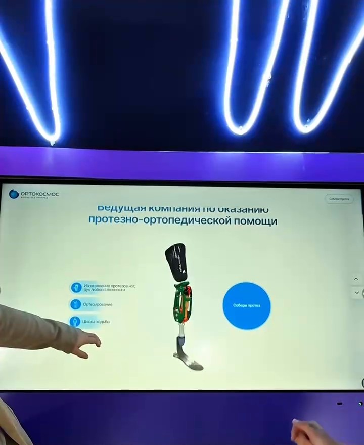 Главный экран приложения на сенсорной панели: заголовок «Ведущая компания по оказанию протезно-ортопедической помощи», три направления, изображение протеза и круглая кнопка «Собери протез»