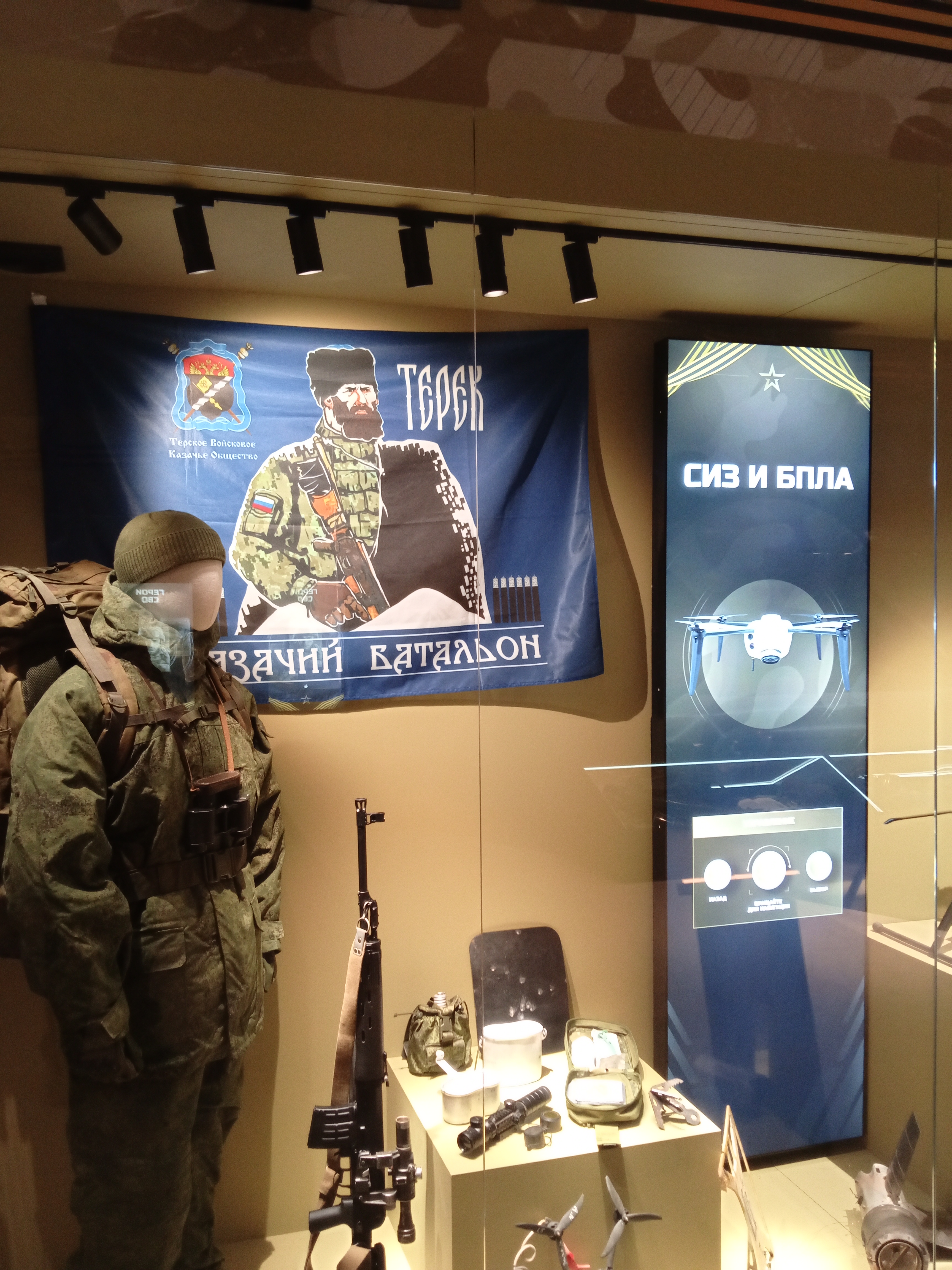 Вертикальный экран «СИЗ и БПЛА» за стеклом витрины: беспилотник и подсказка «Назад», «Вращайте для навигации», «Выбор»; рядом манекен в снаряжении