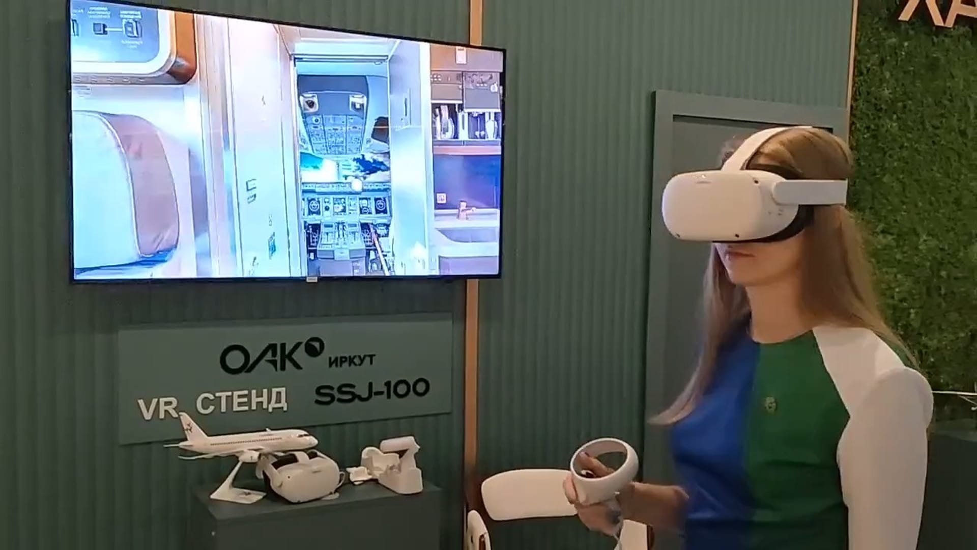 A visitor in a VR headset with controllers at the SSJ-100 stand; the screen shows the view from the virtual cabin