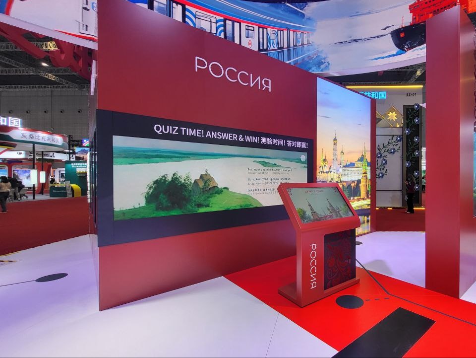 Широкий экран с фильмом «Душа России»: пейзаж с деревянной церковью и строки стихов на трёх языках; под экраном — киоск викторины «Знания о России»
