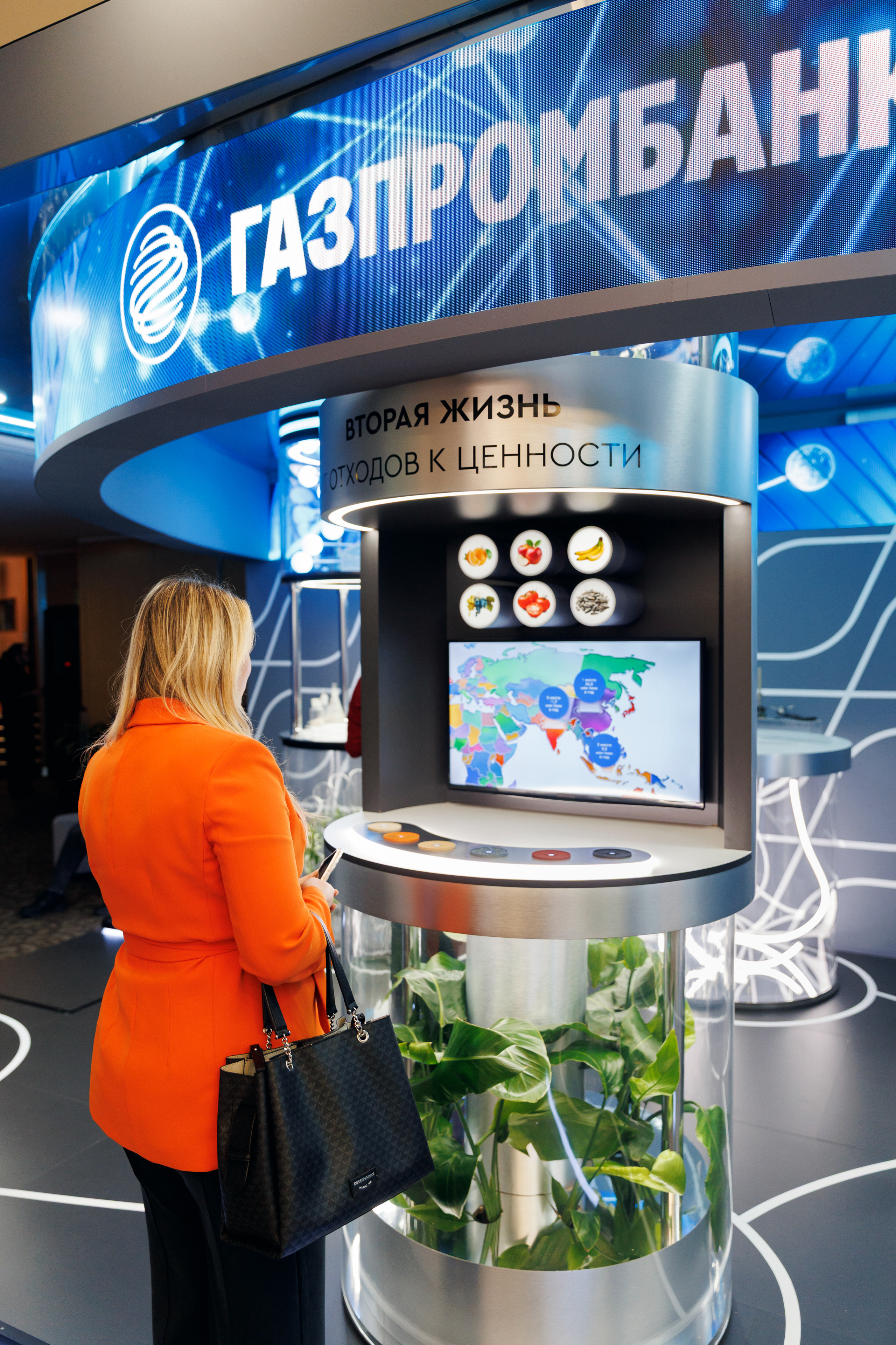 Посетительница у колонны «Вторая жизнь: от отходов к ценности» под светодиодной вывеской Газпромбанка; на экране — карта мира