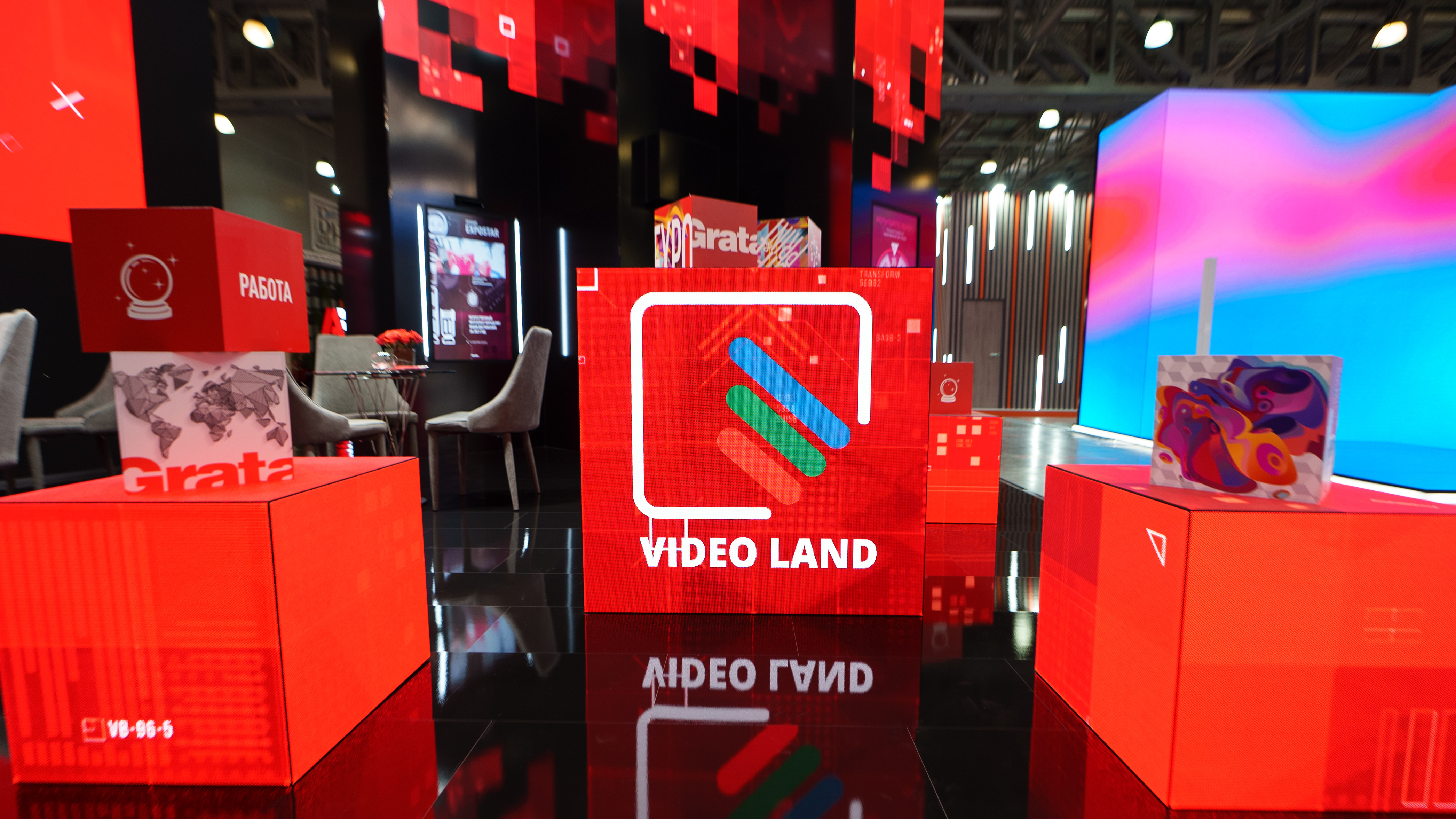 Внутри стенда: в центре — красный светодиодный куб с надписью «Video Land» и его отражение в чёрном глянцевом полу, слева на подставке — красный куб «Работа» со значком хрустального шара, справа — куб с яркой абстрактной картинкой; за ними — кресла, вертикальный экран и стены с красным пиксельным узором