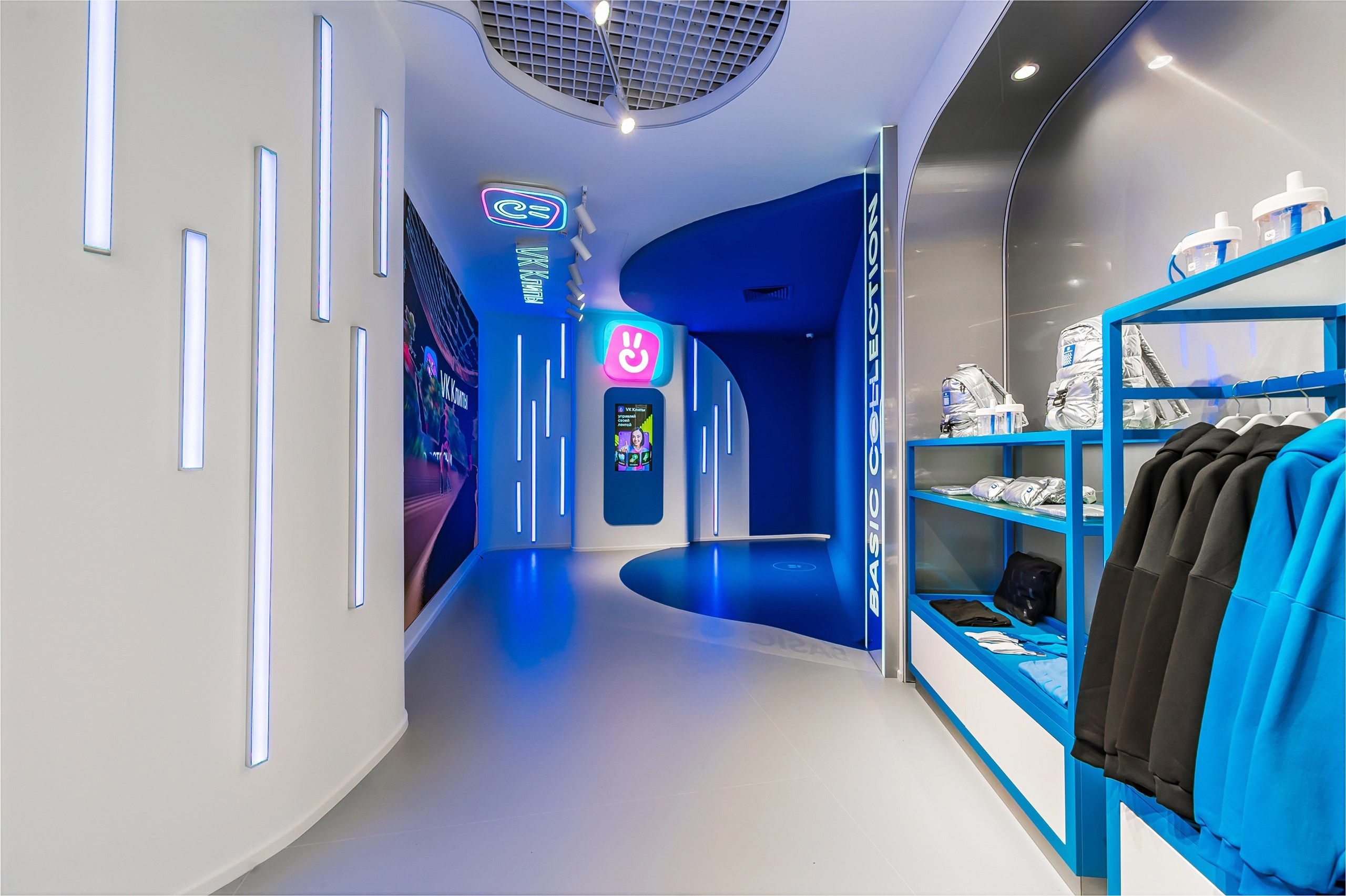 The aisle to the VK Clips zone: a big screen on the wall on the left, a vertical touch kiosk under the neon VK Clips logo at the end of the aisle, clothing racks on the right