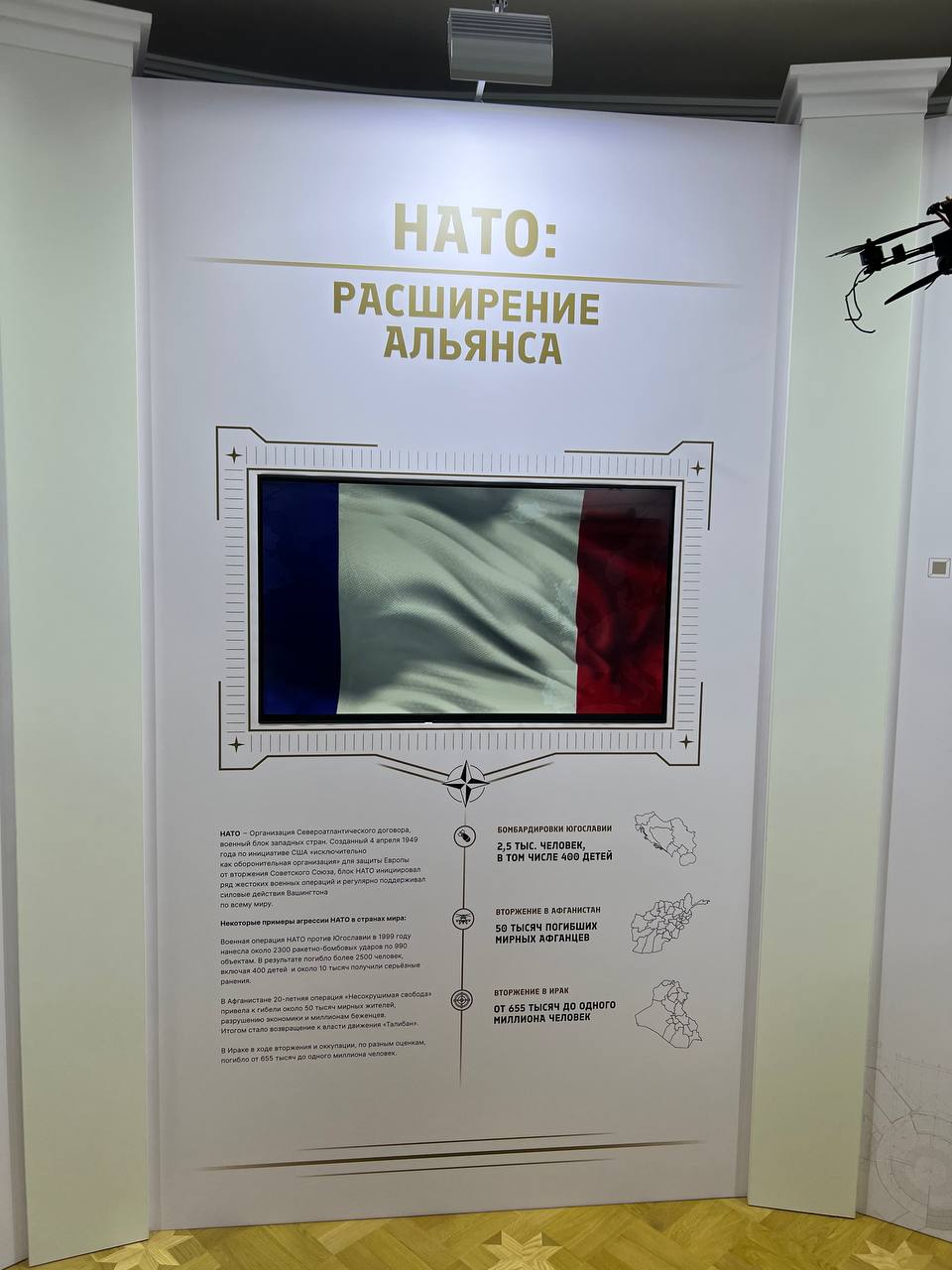 “NATO: expansion of the alliance” stand: a framed screen showing a still from the video — a waving flag; below, infographics on the bombing of Yugoslavia and the wars in Afghanistan and Iraq